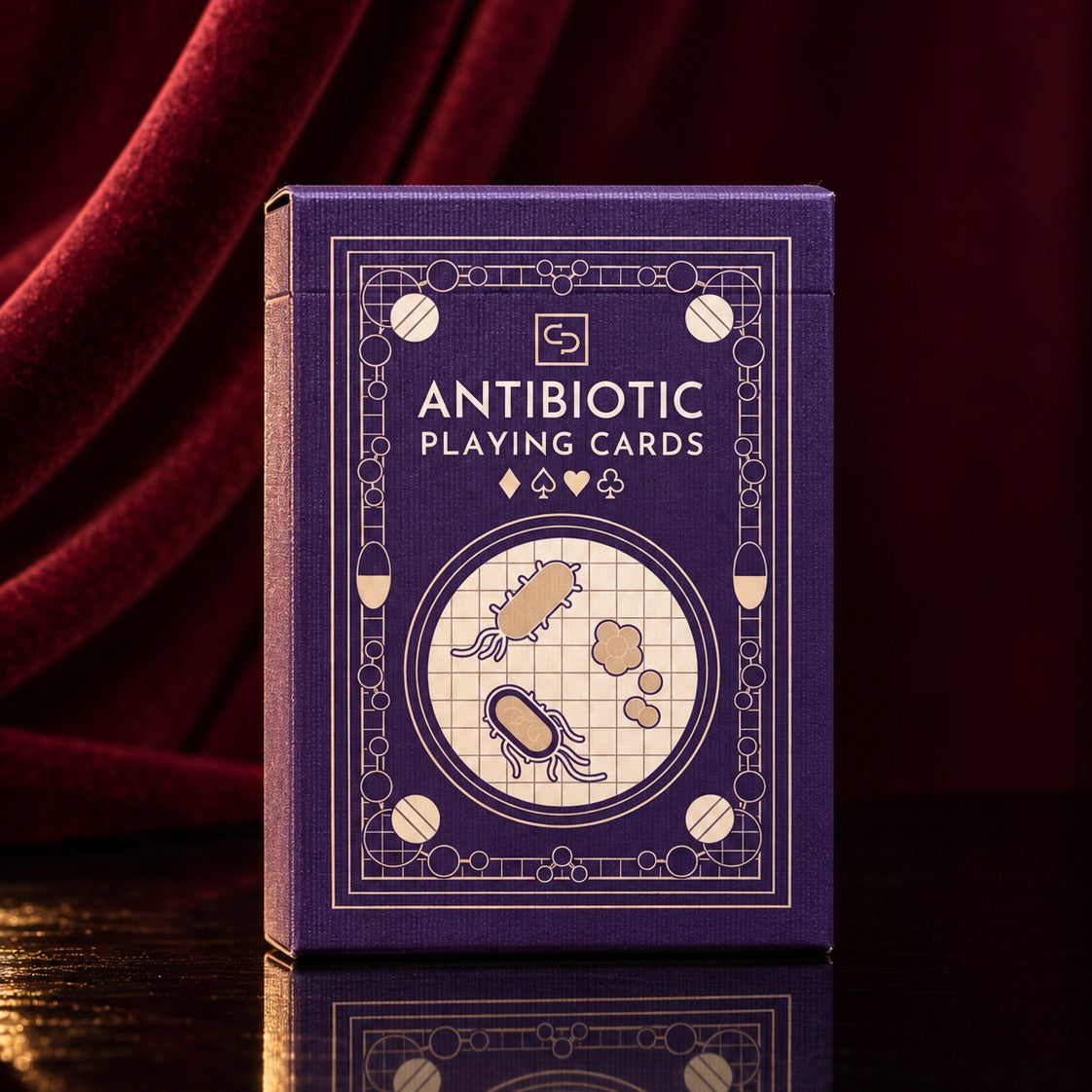 The Antibiotic Playing Cards tuck box, gold-foiled on purple, against red velvet